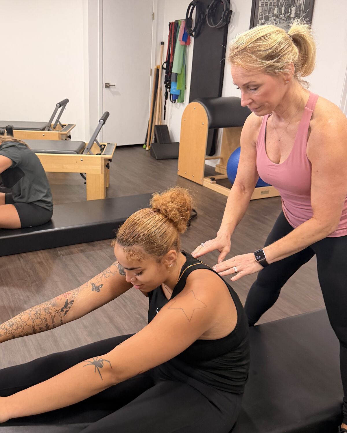 A coach guiding a client’s back as she reaches for the bar in a seated stretch on the reformer